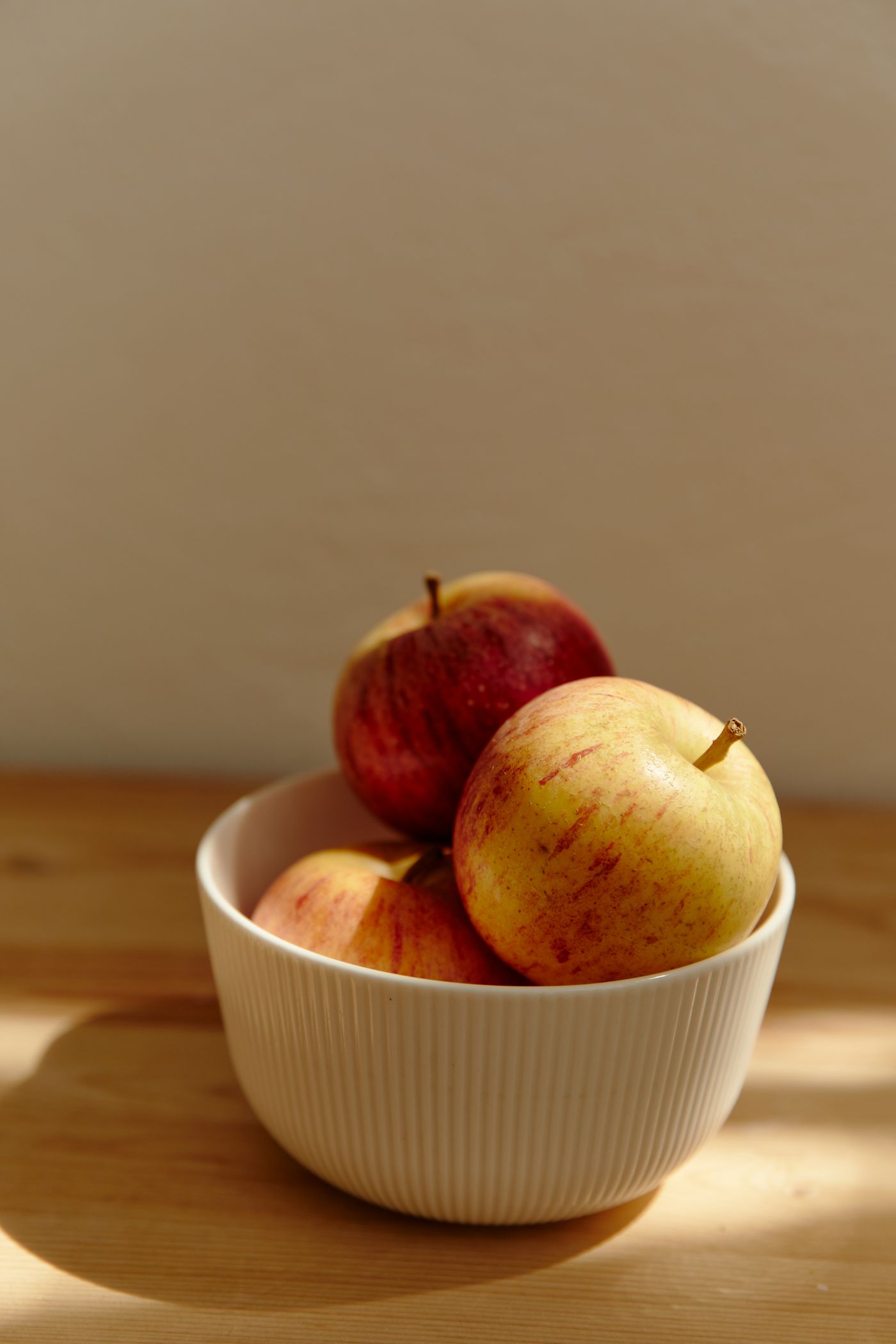 Three red-and-yellow apples in a cream ribbed ceramic bowl on light wood, with a beige wall behind.