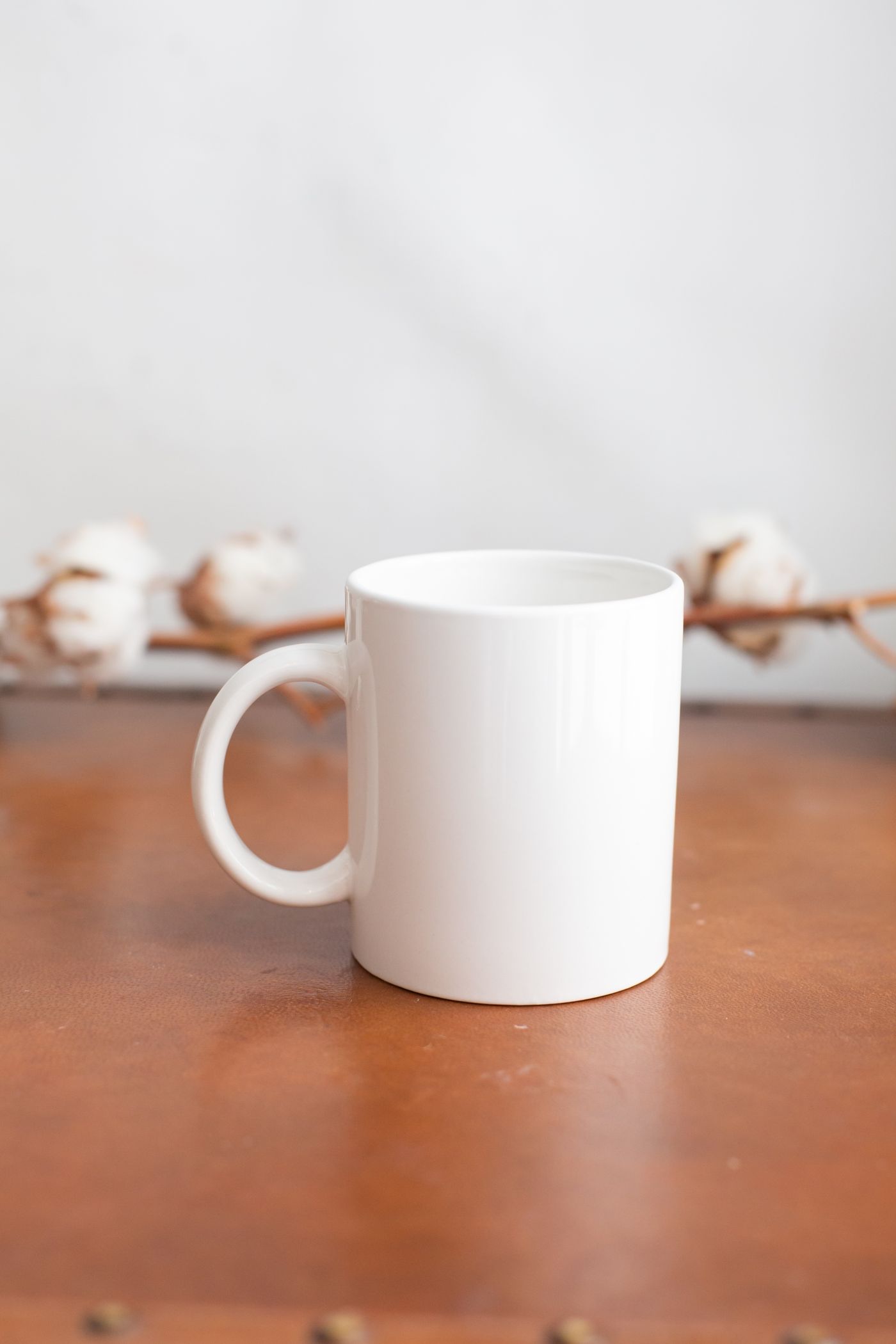 A plain white glossy mug on a rust-orange leather-look surface with brass studs along the bottom edge, with a branch of white cotton bolls behind it against a pale grey wall.